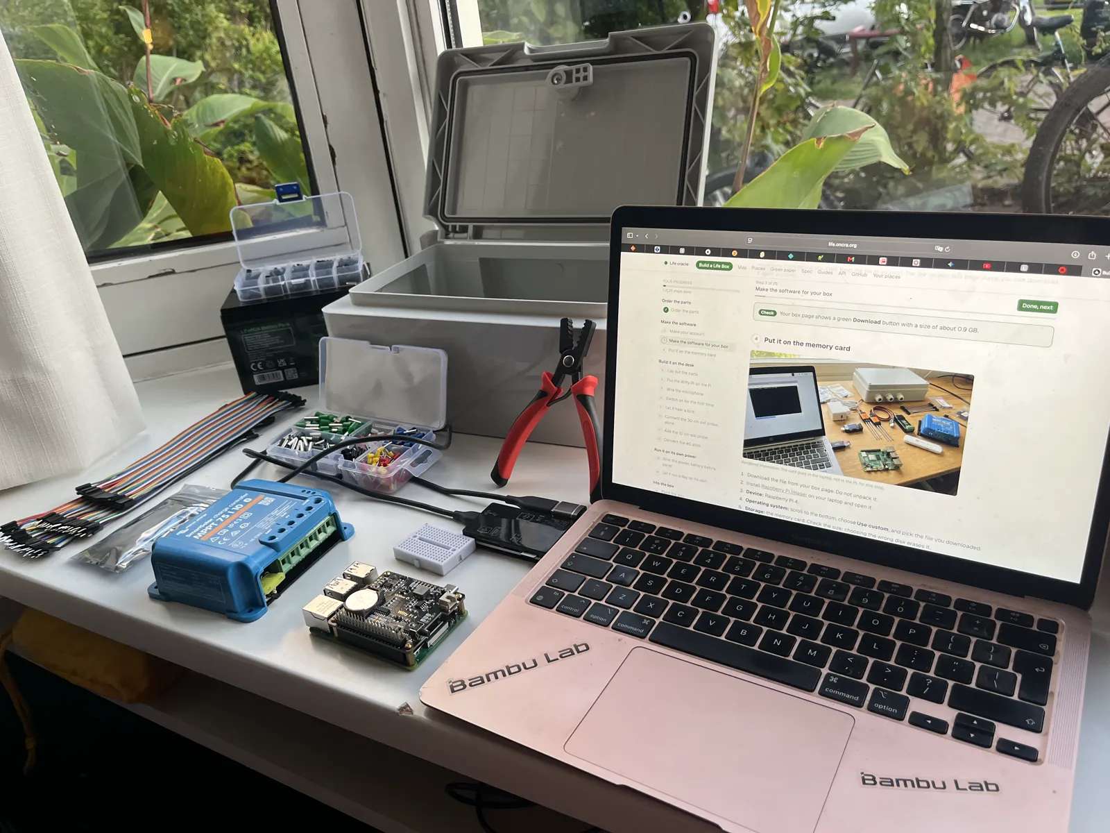 The build table at the window: the open enclosure, the black battery, the blue charge controller, the Raspberry Pi with the Witty Pi, ferrules and pliers, and the laptop with this guide open