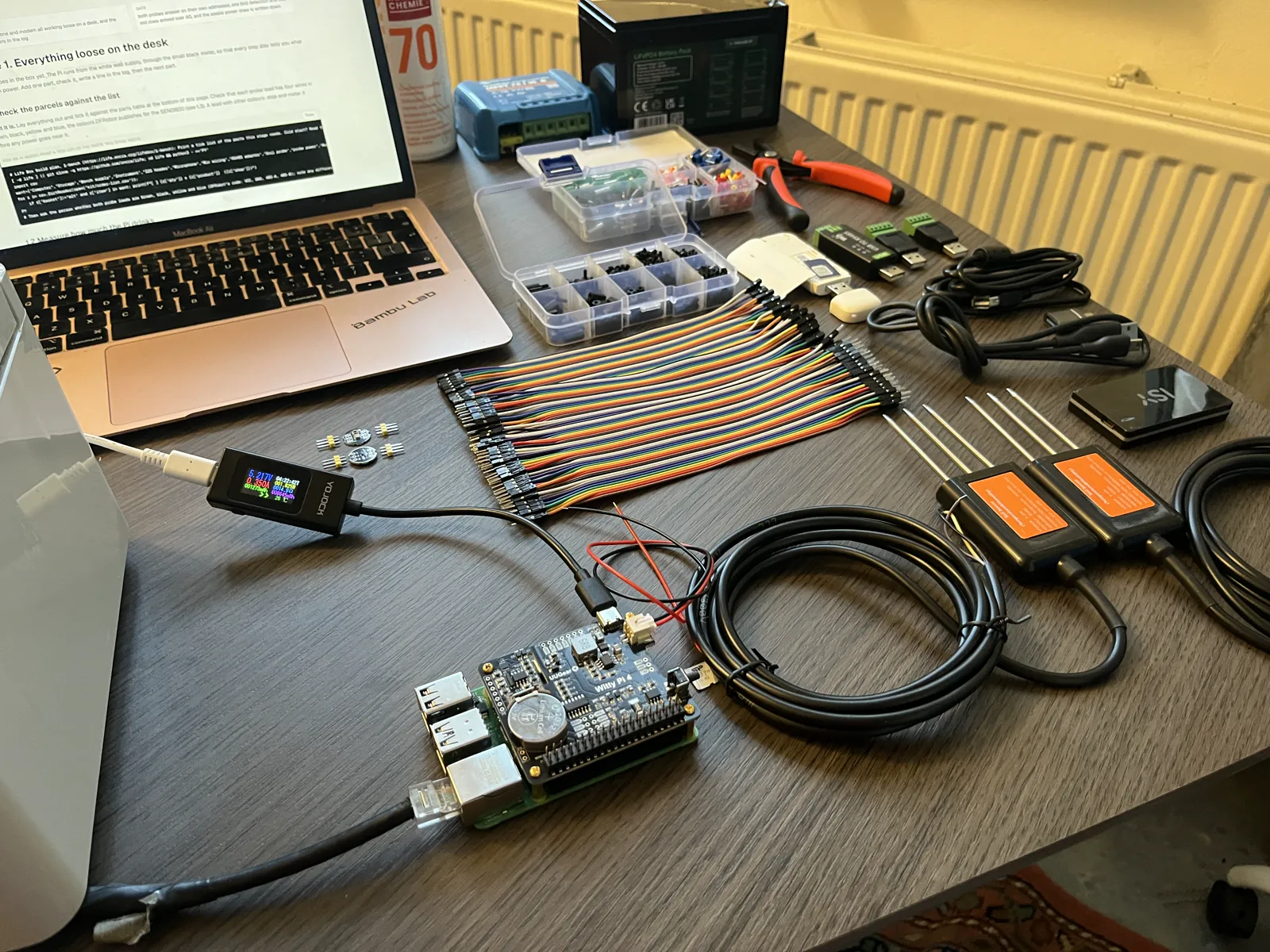 The desk with the Raspberry Pi and Witty Pi in front, the meter in the cable from the wall supply, soil probes, jumper wires and parts boxes behind