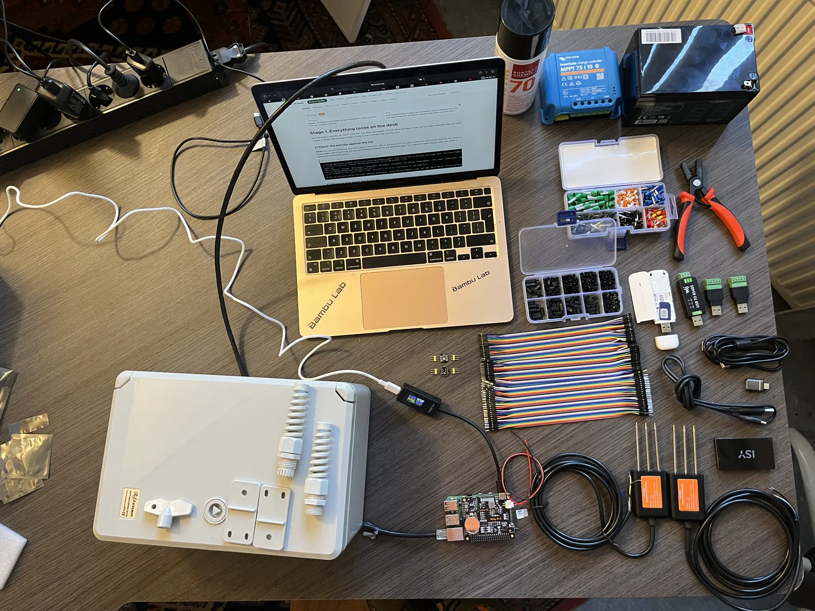 All the parts for one box laid out on a table, seen from above: enclosure with glands, laptop, Raspberry Pi with Witty Pi, meter, microphones, jumper wires, standoffs, ferrules, 4G stick, RS485 adapter, USB screw terminals, crimping pliers, two soil probes, charge controller, battery