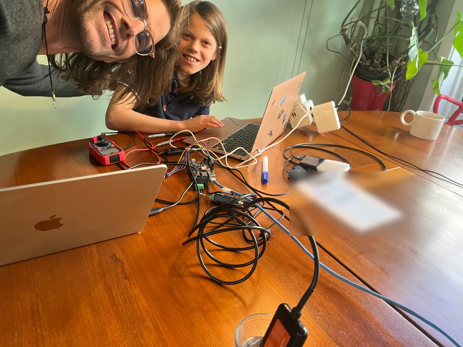 Two of the builders leaning into the picture at the desk, laughing; in front of them two laptops, a multimeter, the Raspberry Pi with the Witty Pi and its coloured microphone wires, and a coil of probe cable