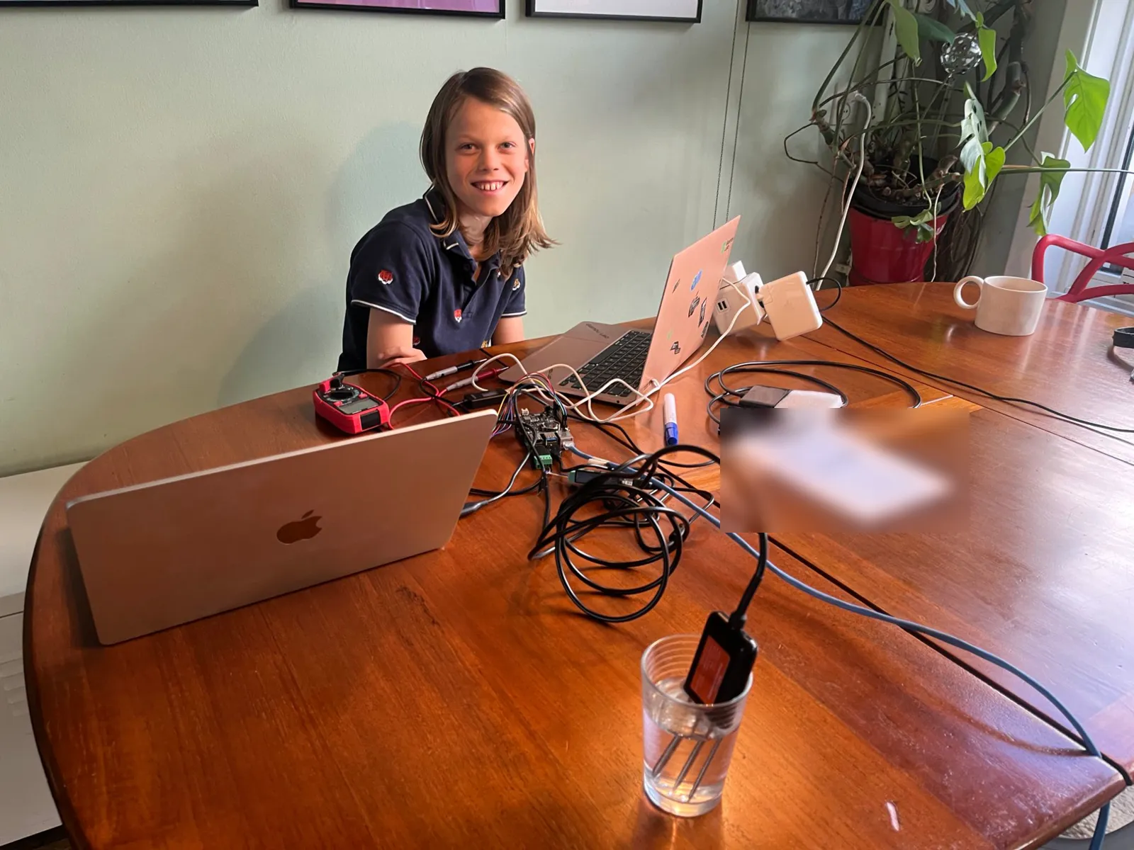 A smiling builder at the desk behind two laptops and a multimeter; in the foreground the soil probe stands in a glass of water, its black lead curling back to the Raspberry Pi
