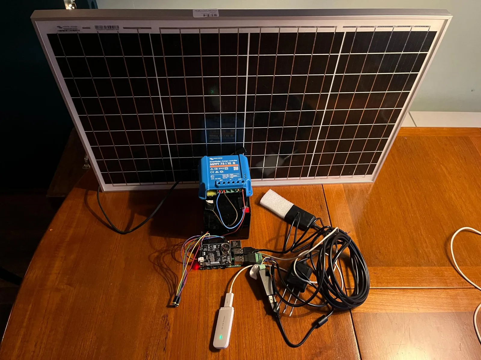 The full chain back on the table afterwards: the panel propped upright, the blue Victron charge controller wired to the battery, the Witty Pi and RS485 adapter in their tangle of coloured wires, and the 4G stick's green light in front