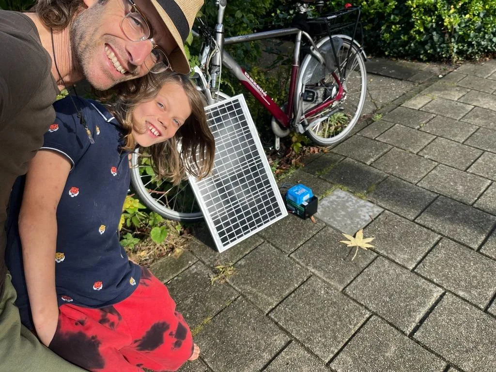 One of node 1's builders and its youngest, crouched beside a parked bicycle on a paved path, both smiling; the solar panel leans against the bike's back wheel, its lead trailing to the battery case on the ground
